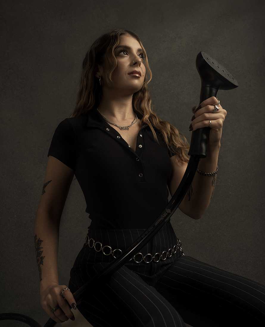 Woman with long wavy hair holds a black shower head, wearing a black shirt and pinstriped pants, against a dark gray background.