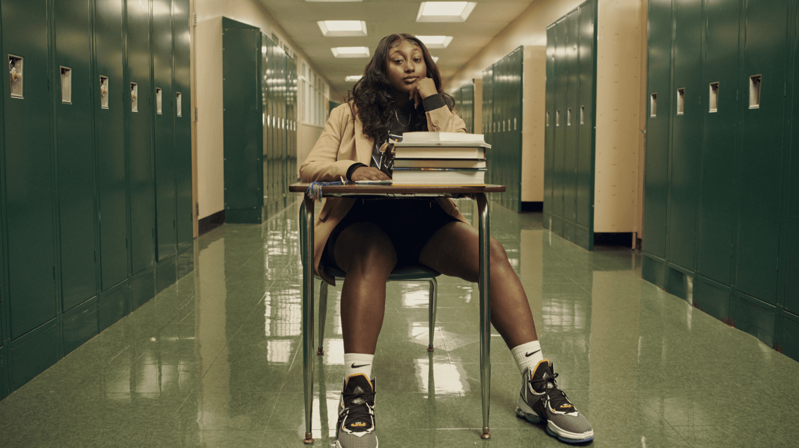 Girl sitting at school desk that has school books stacked on it. Desk is in a school hallway with lockers lining the walls. Girl is wearing Nike LeBron XIX shoes