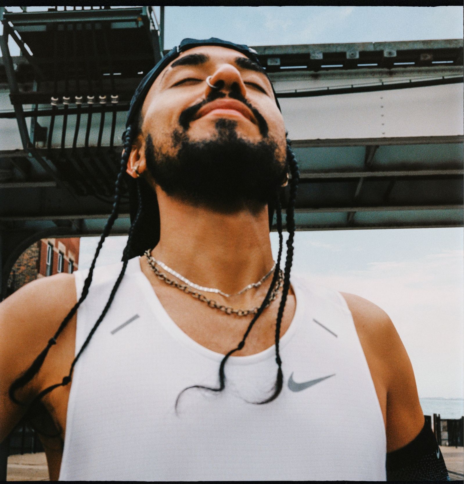 Man with nose ring, beard, braided hair and white Nike top has head tilted up, eyes closed and smiling