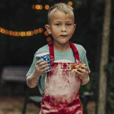 A young boy with flour on his face and red apron holds a paper cup and a slice of pizza outdoors at night; string lights are visible in the background.