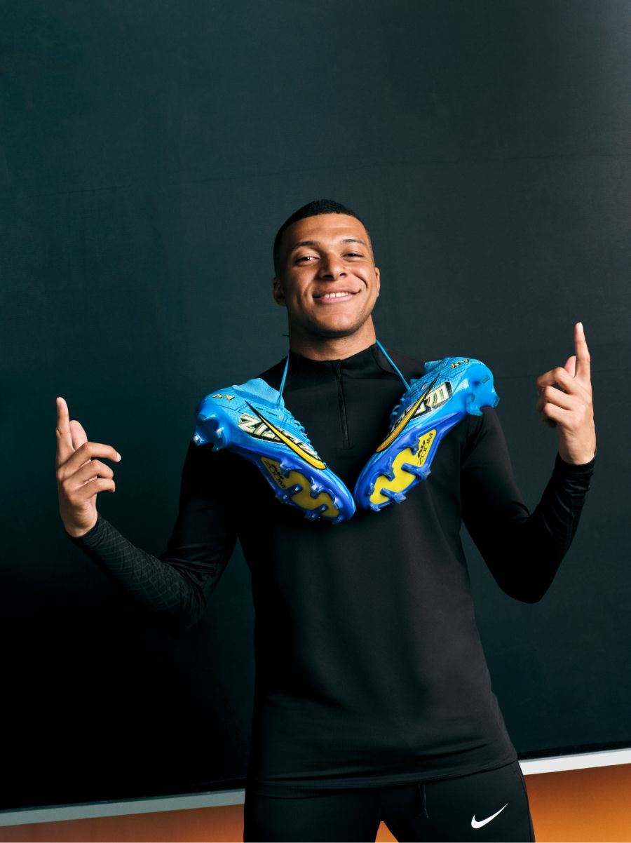Mbappe with Nike Mercurial boots around his neck, pointing up and smiling