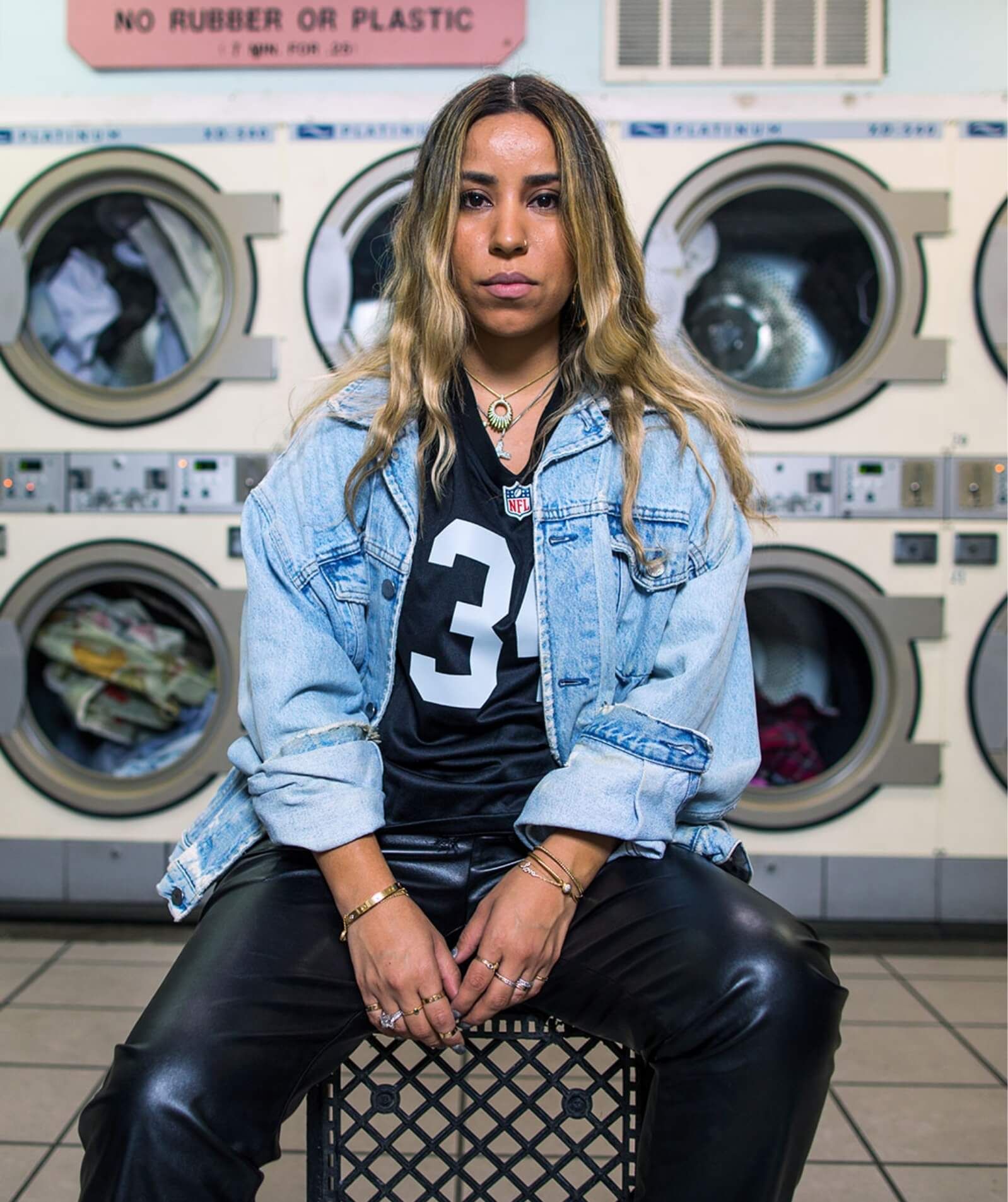 Woman wearing denim jacket and number 34 Raiders jersey in laundromat sitting on crate