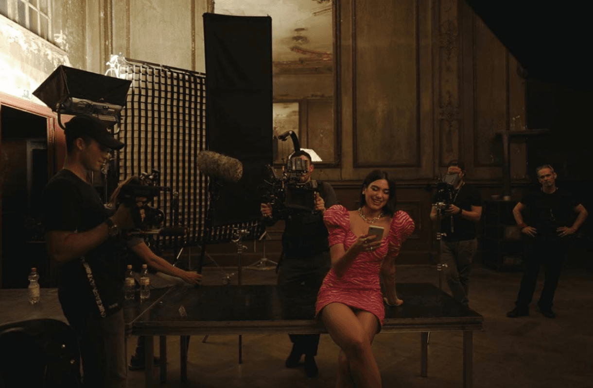 Behind the scenes Karol G video shoot. She is wearing pink dress and looking at her phone surrounded by camera equipment and crew