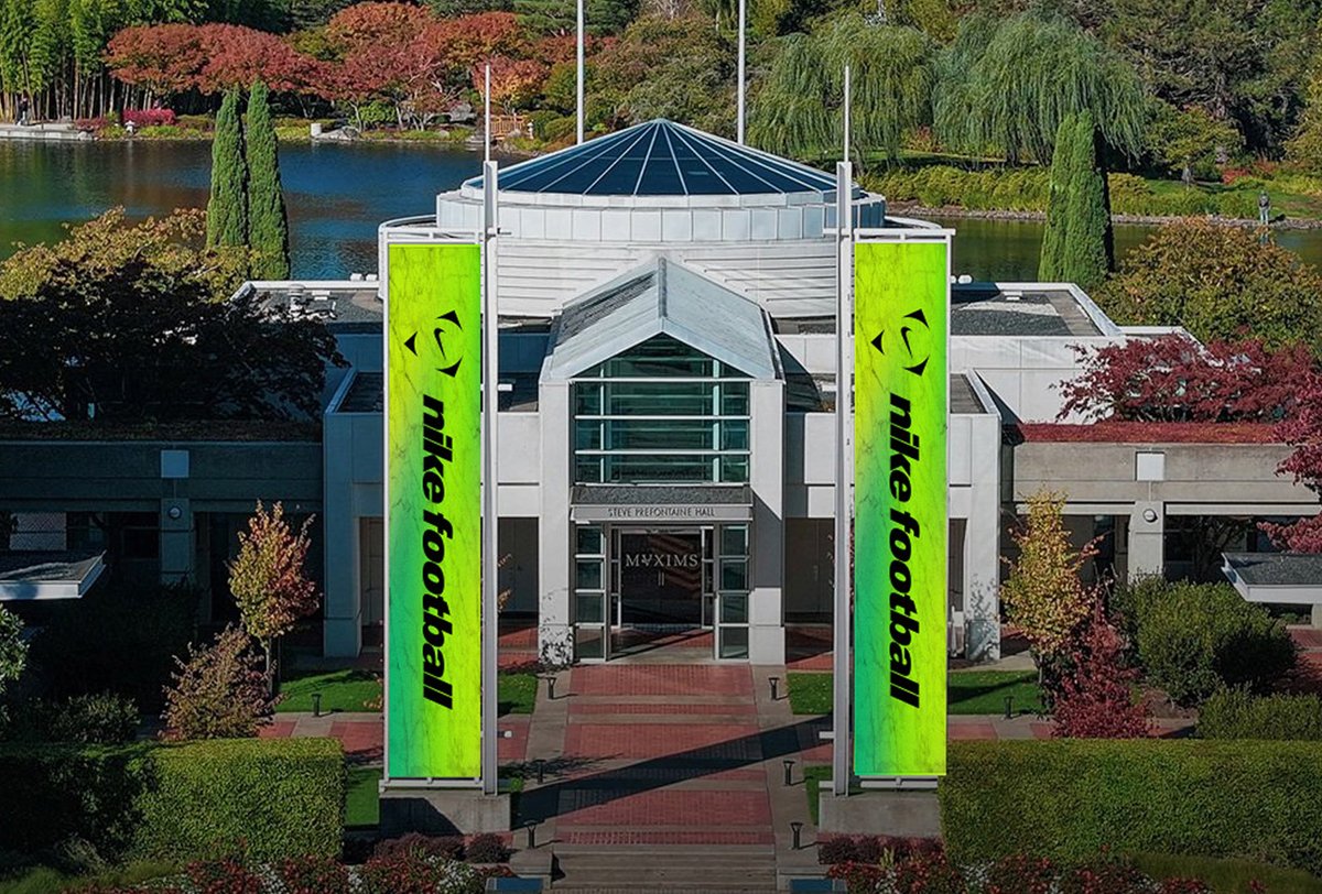 A glass and stone building with two tall, bright green banners reading "nike football" stands amid landscaped trees and a lake in the background.