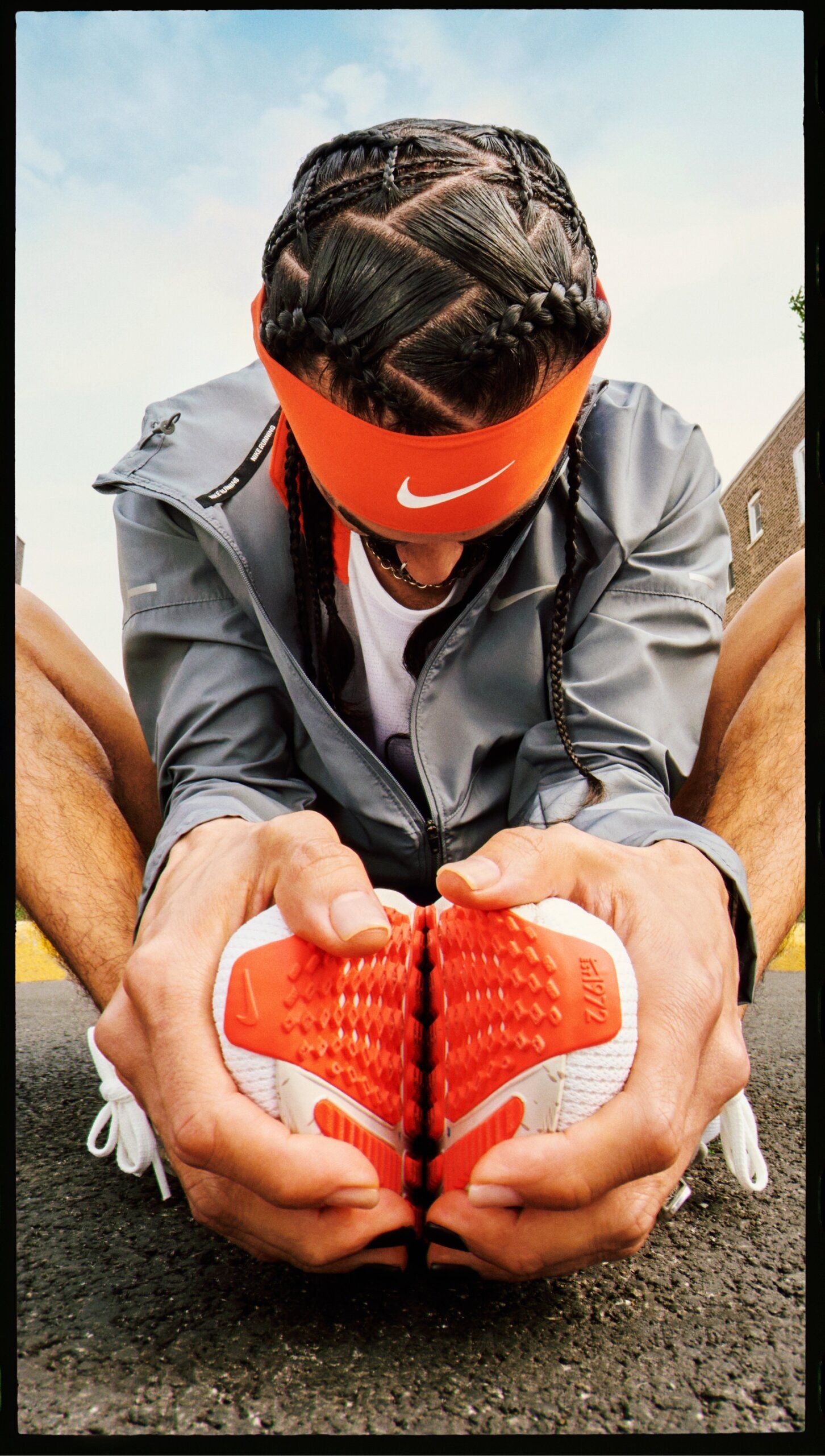 Man stretching in street in cobblers pose head down. He has geometric, parting and braiding in hair with orange nike headband.