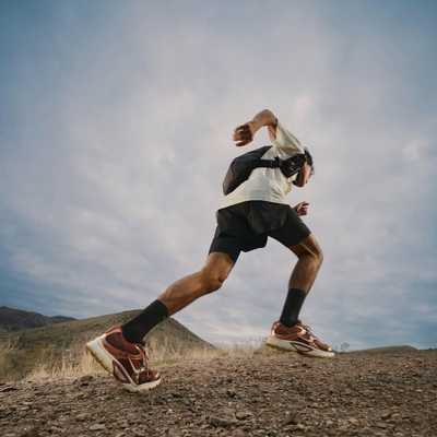 A person wearing athletic gear and a backpack runs uphill on a rocky trail with mountains and a cloudy sky in the background.