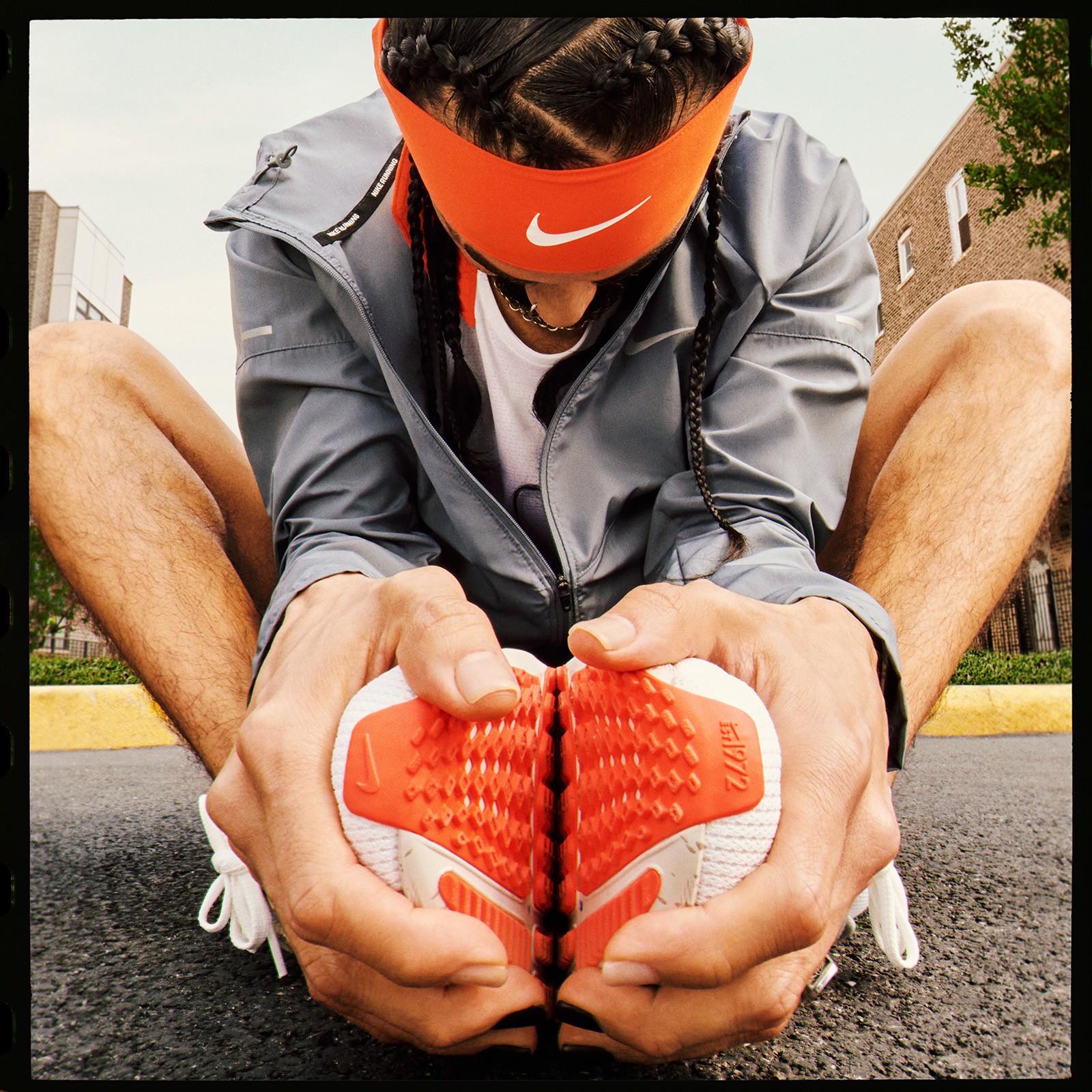 Man stretching in street in cobblers pose head down. He has geometric, parting and braiding in hair with orange nike headband.
