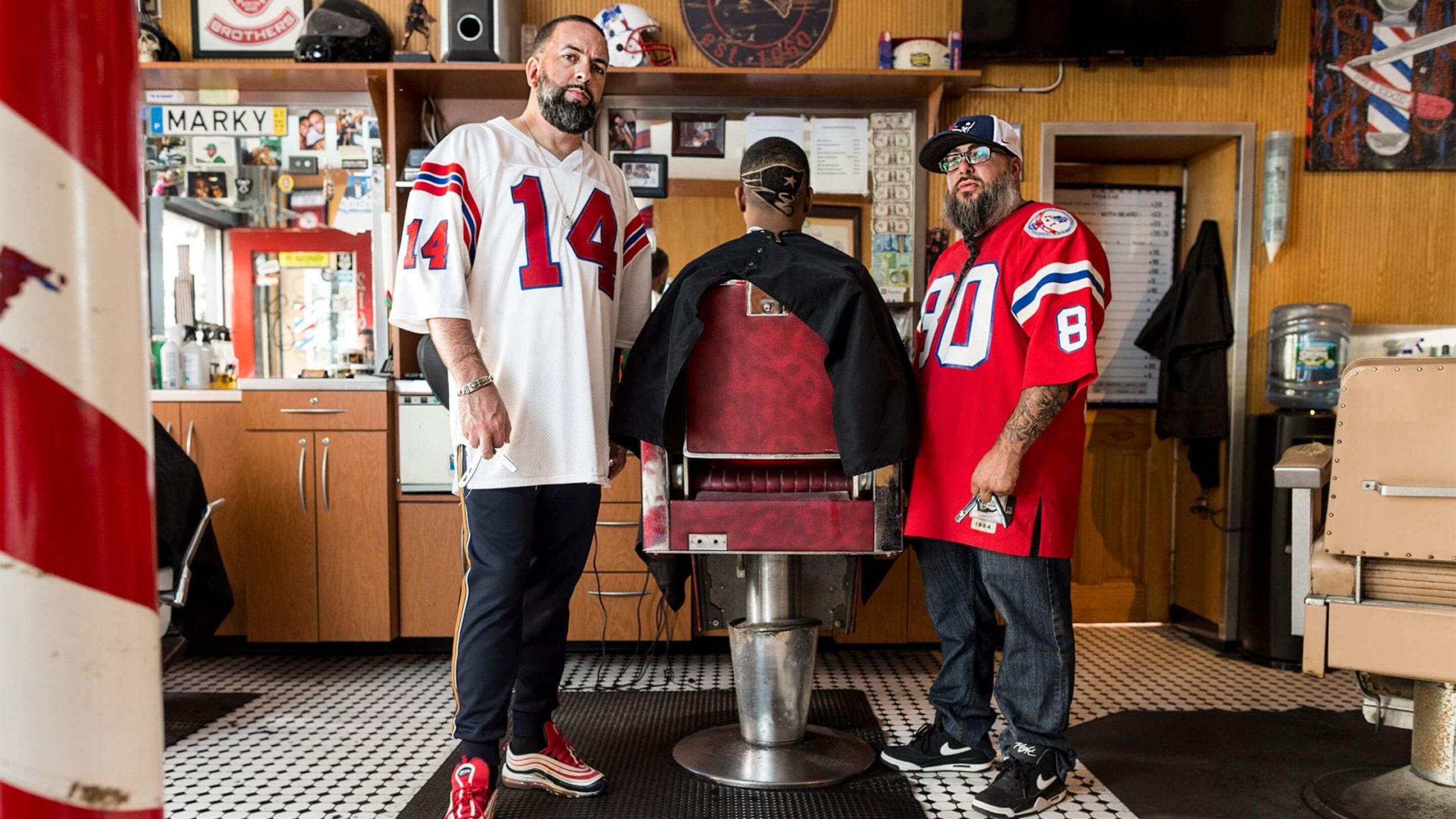 New England Patriots logo shaved into back of head of man sitting in barber stool with two barbers in Patriots Jerseys next to him