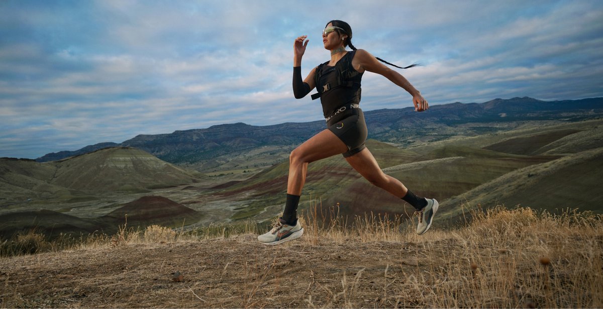 A person wearing athletic gear runs on a dirt trail with hills and mountains in the background under a cloudy sky.