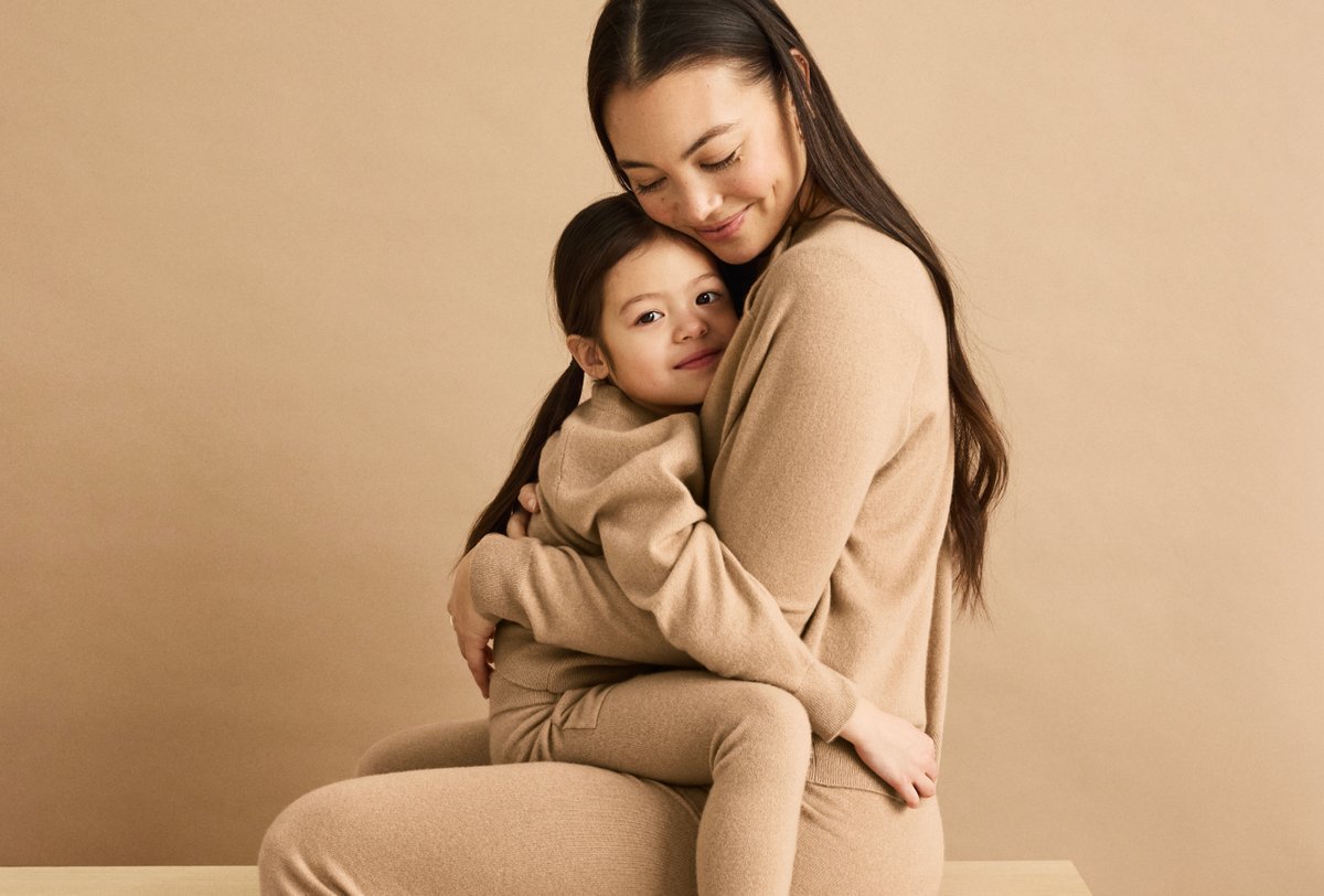 A woman sits and embraces a young child, both dressed in matching beige clothing, against a beige background.