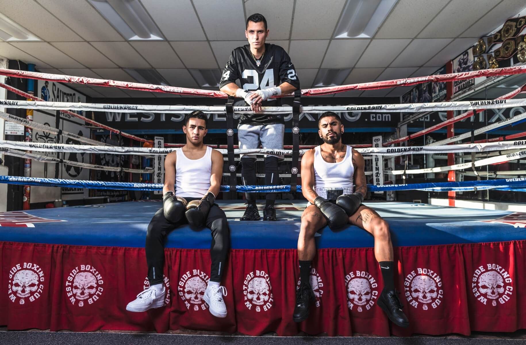 Three men resting against ropes in boxing gym, one has number 24 Raiders football jersey on